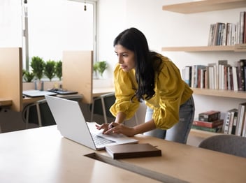Woman in a yellow blouse stands and types on a laptop at a desk in a modern office with bookshelves and potted plants in the background.
